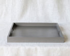 Front-facing view of a rectangular grey ottoman tray with handles on each side, set against a light fabric background.