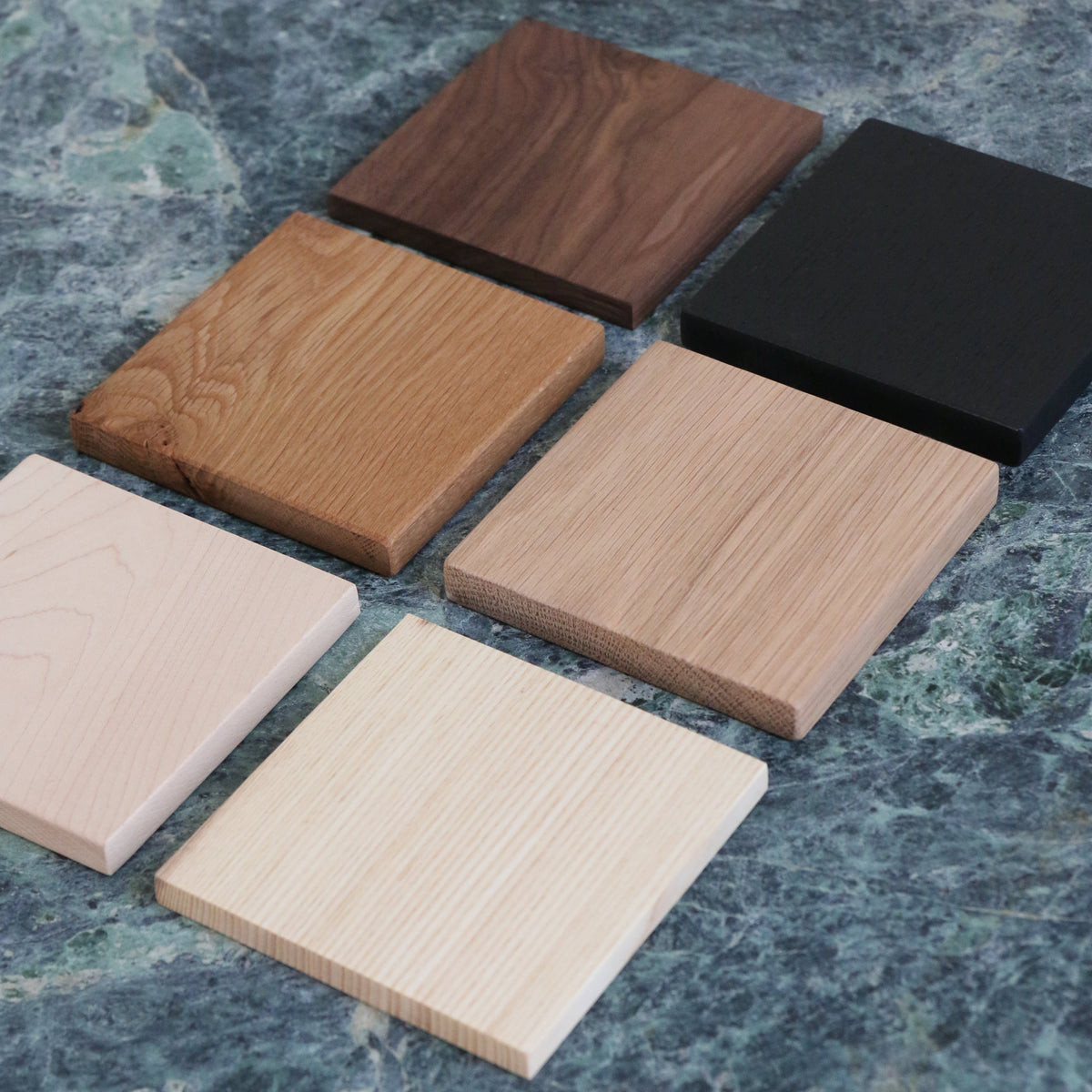Six square wood swatches in different natural wood tones and one black wood swatch, arranged on a mottled green stone surface.