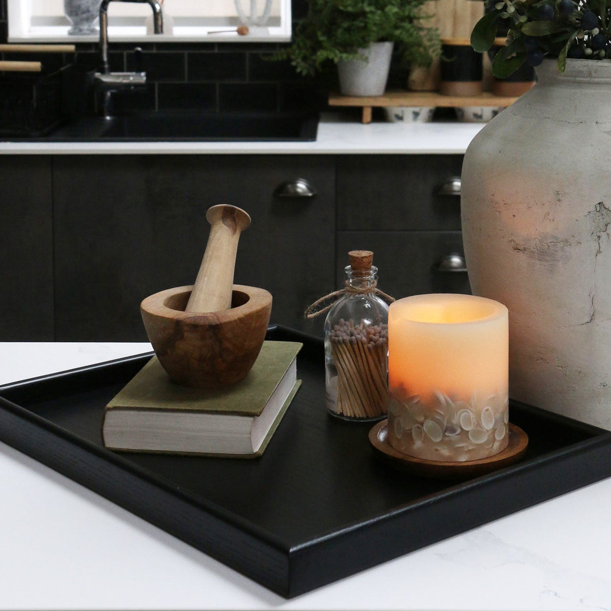 A decorative tray on a kitchen counter, styled with a candle, a book, and a wooden mortar and pestle, showcasing its versatility.