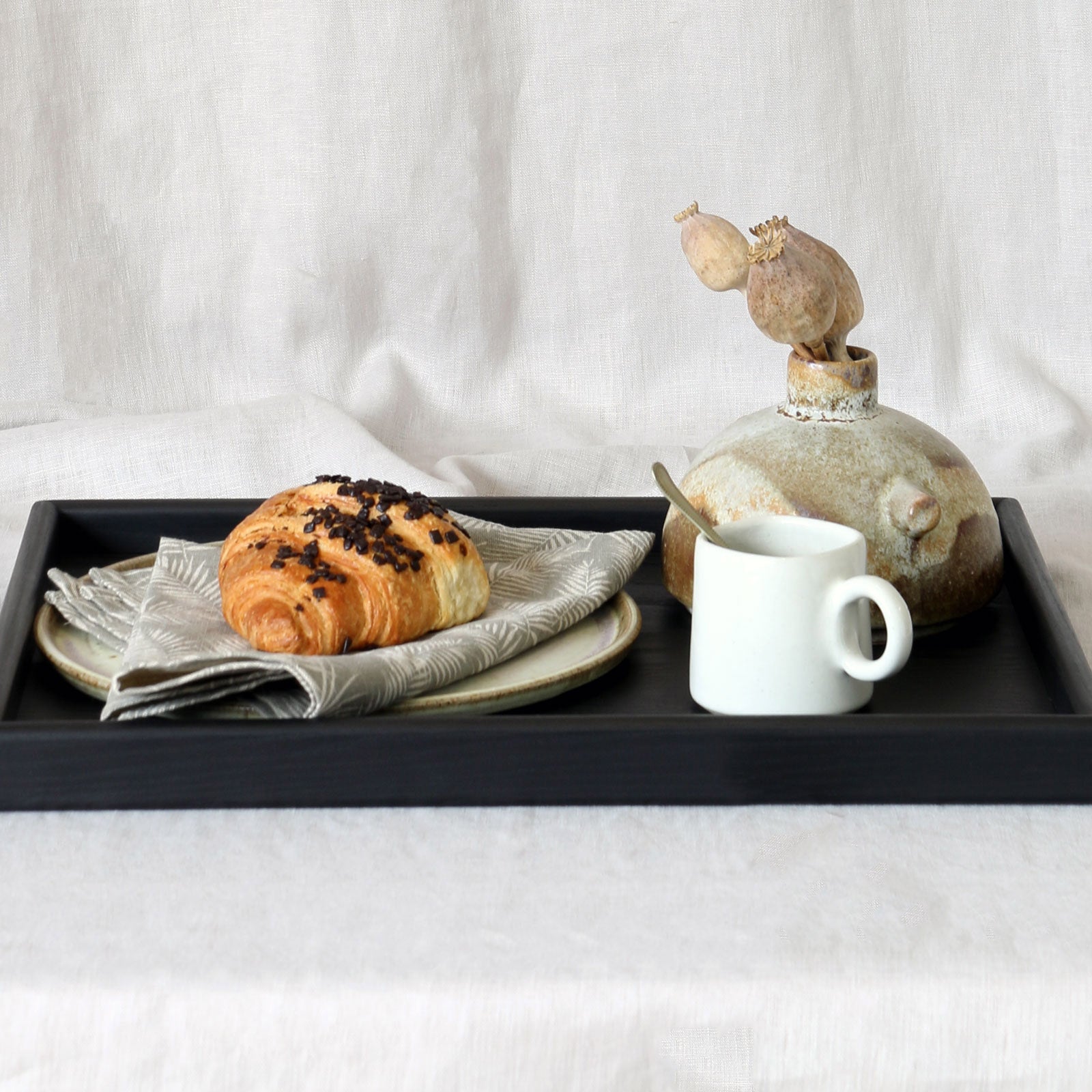 Wooden tray for footstool featuring a black handcrafted design, elegantly styled with a breakfast setting including a croissant, a white ceramic cup, and a rustic decorative vase.