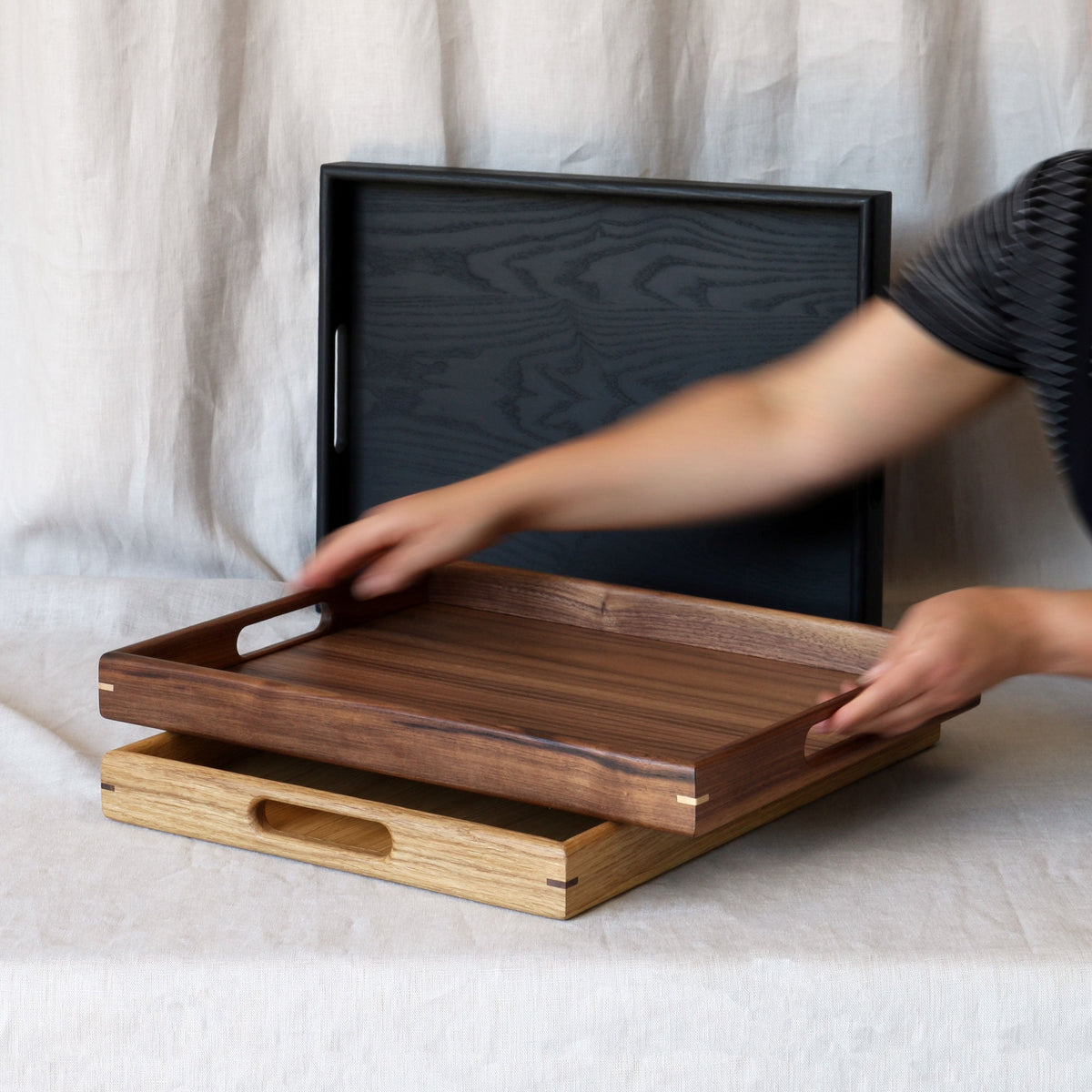 A person's hands and arms are shown holding a medium-brown walnut serving tray with a handle, resting on top of a light oak wood tray. A black wooden tray with a handle stands upright in the background against a draped beige fabric backdrop.
