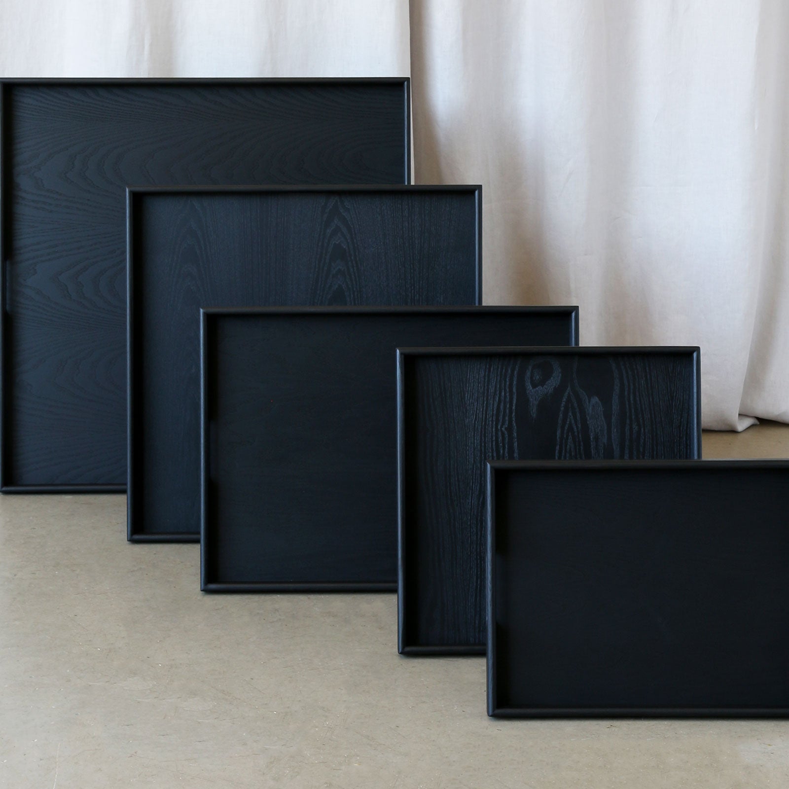 A set of handcrafted trays in black wood finish, neatly stacked in descending sizes against a neutral backdrop.