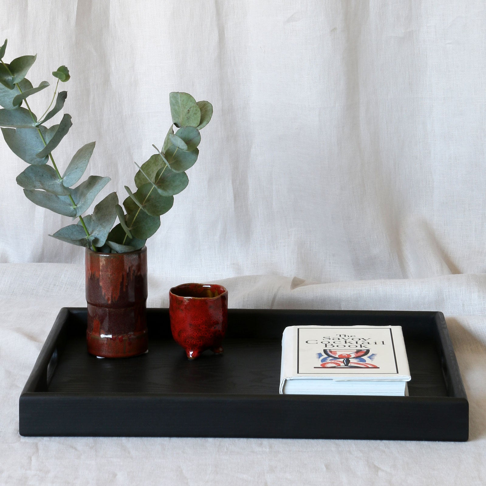A decorative tray in black, adorned with eucalyptus stems in a ceramic vase, a red cup, and a book, displayed on a soft fabric surface.