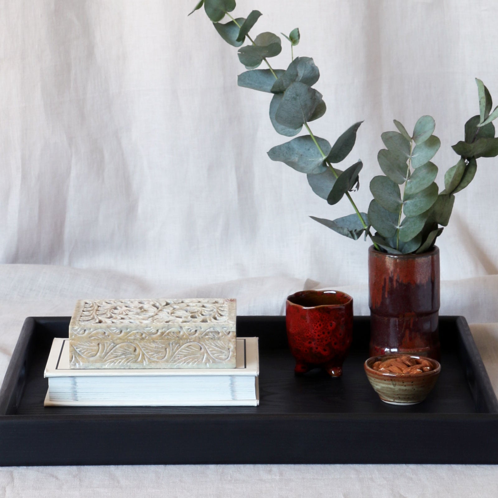A decorative tray in black with books, a ceramic vase with eucalyptus, and artisanal pottery cups, styled elegantly.