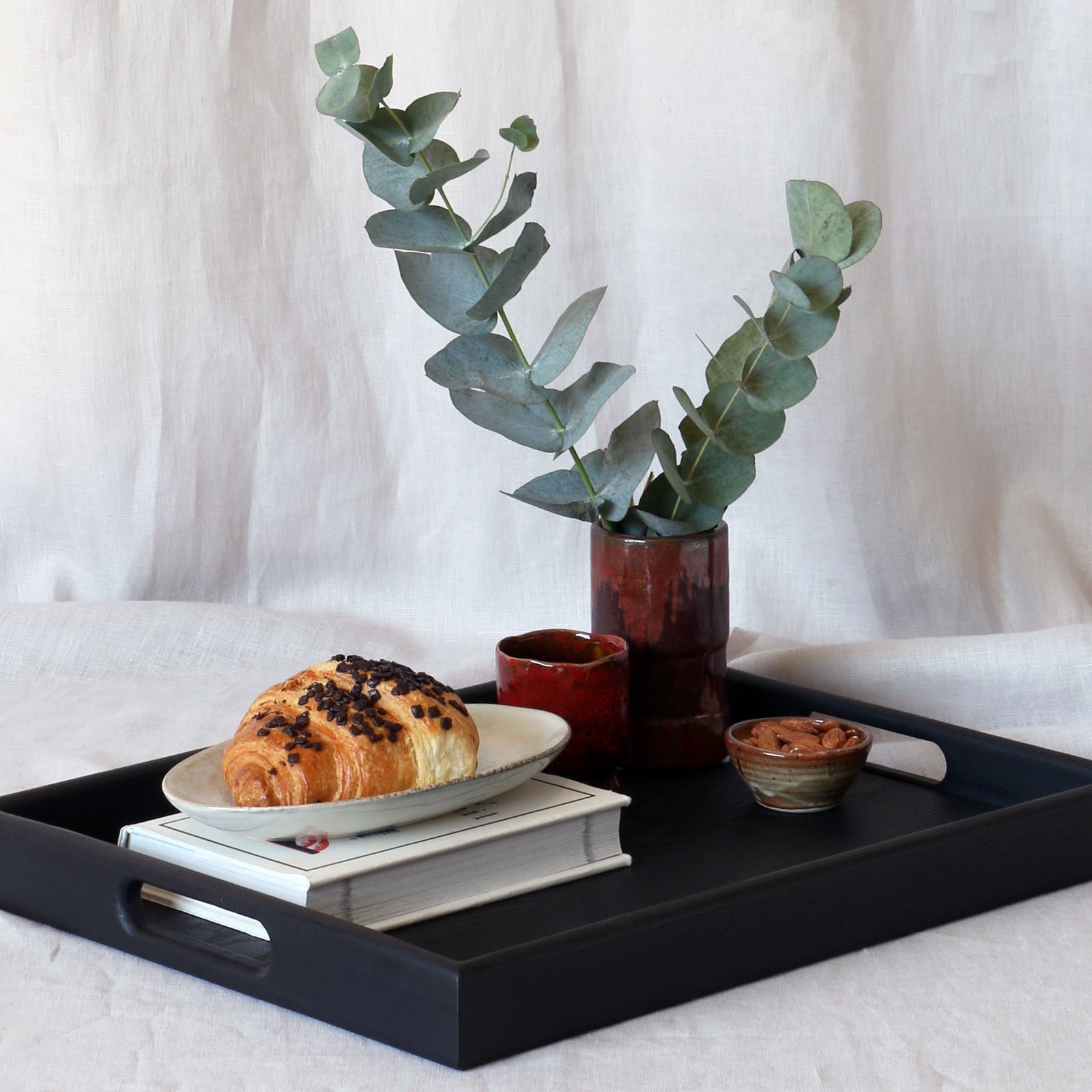 A coffee table tray in black holding a plate with a chocolate croissant, a stack of books, and decorative ceramic items, styled with eucalyptus stems.