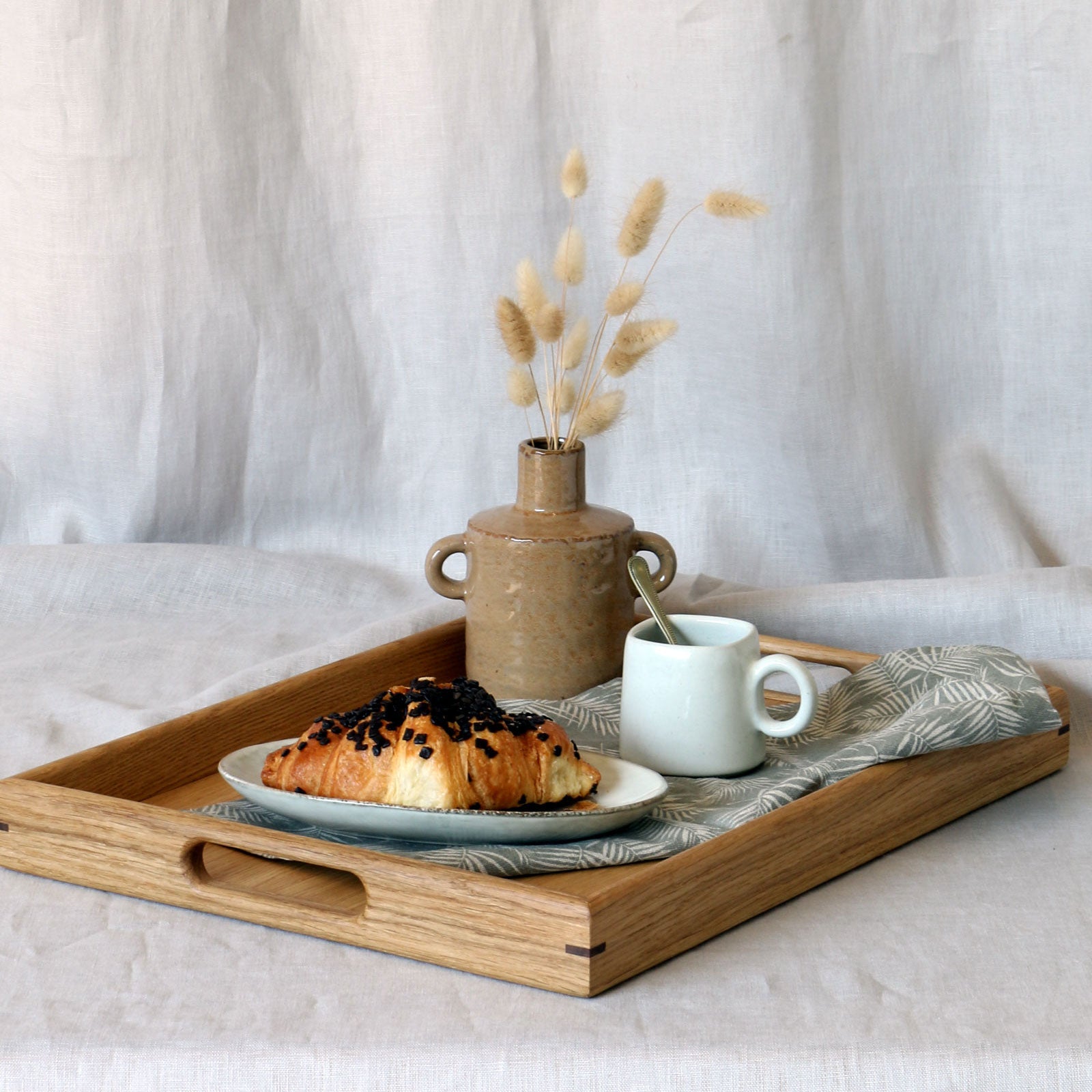 Breakfast-in-bed tray with handles, featuring a rustic ceramic vase, a cup of coffee, and a chocolate croissant on a linen napkin, set against a neutral fabric backdrop.