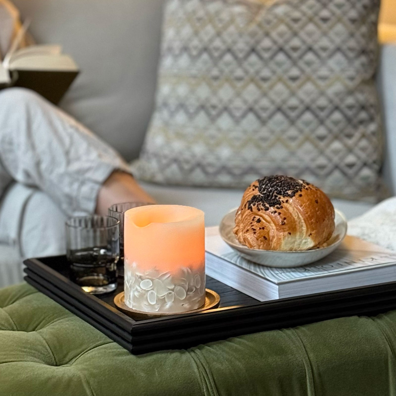 A wooden tray for a footstool, featuring a handcrafted black wood design with reeded edges, styled with a candle, glasses, and a croissant, creating a cozy and elegant living space.