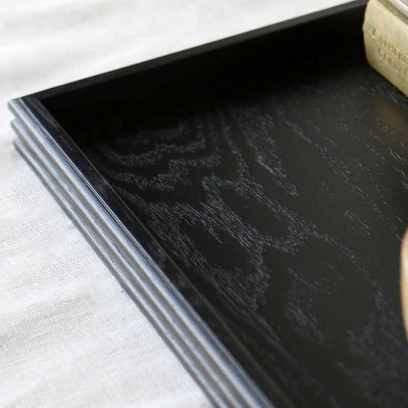 Close-up of a black wooden tray corner, showing the rich wood grain texture and reeded detailing along the edges, partially styled with a ceramic cup and book.