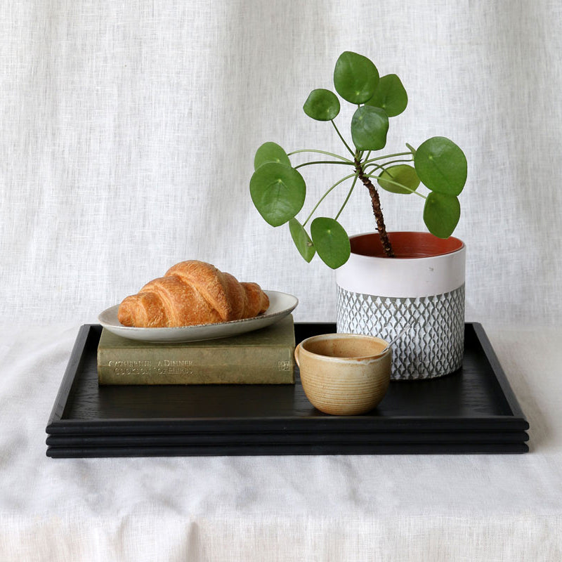 The black tray styled with a plate of croissants, a ceramic mug, a potted Pilea plant, and a hardcover book, placed on a white fabric surface.