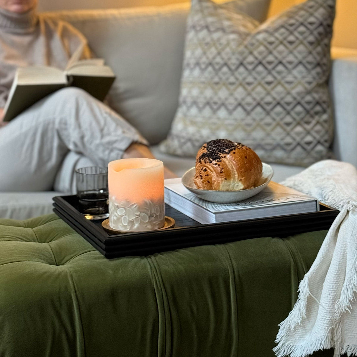 A cosy living room scene with a woman sitting on a light grey sofa reading a book, while a green velvet ottoman in the foreground holds a black decorative tray with a candle, a book, and a pastry. A patterned throw pillow, a lit table lamp, and a white throw blanket are visible in the background.