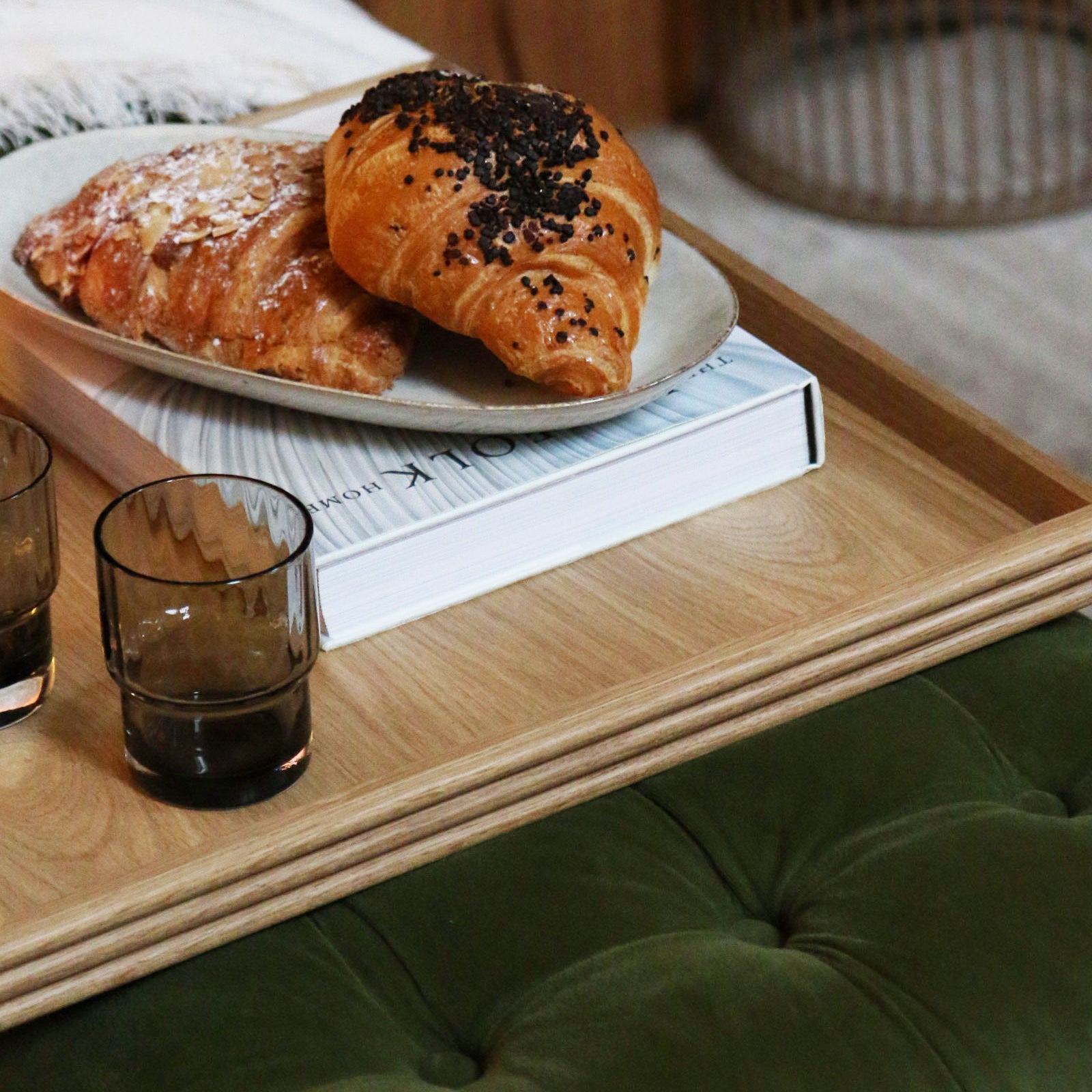 A coffee table tray in oak, neatly placed on a green tufted footstool, holding glasses, a croissant, and a book, adding warmth and functionality to the space.