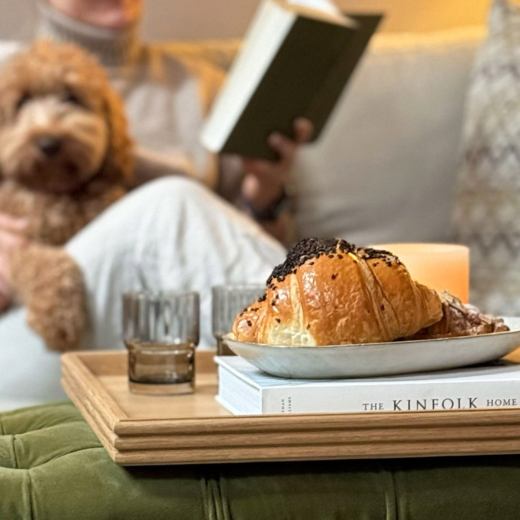A handcrafted tray in natural oak, beautifully displayed with pastries, a candle, and glasses, set on a tufted ottoman, enhancing the ambiance of a cozy reading nook.