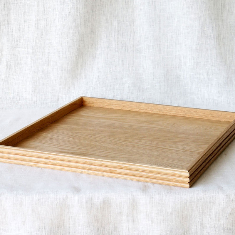 A square wooden tray made from oak, featuring subtle reeded detailing along the edges, placed on a soft white linen backdrop.