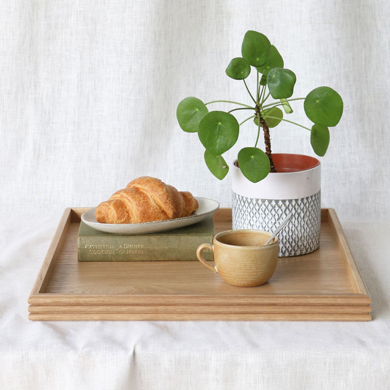 The oak tray styled with a croissant on a plate, a ceramic mug with a spoon, a potted Pilea plant, and a hardcover book, all resting on a white fabric surface.