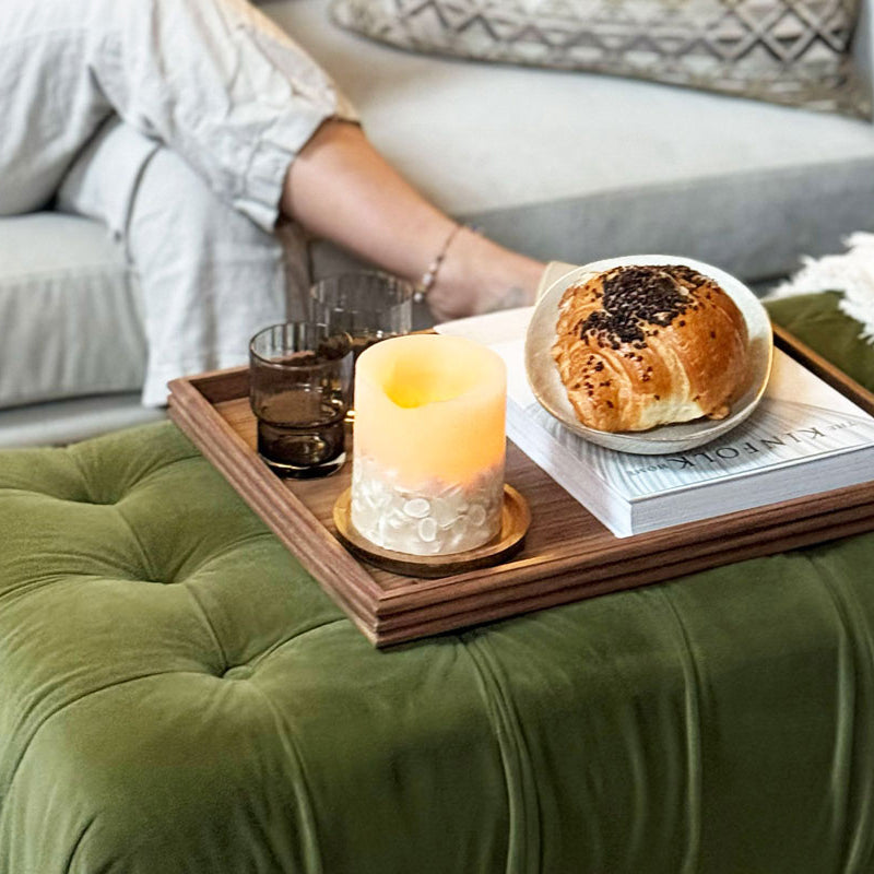 A decorative tray made of walnut, placed on a tufted ottoman, holding a croissant, book, and candle, creating a warm and inviting aesthetic.