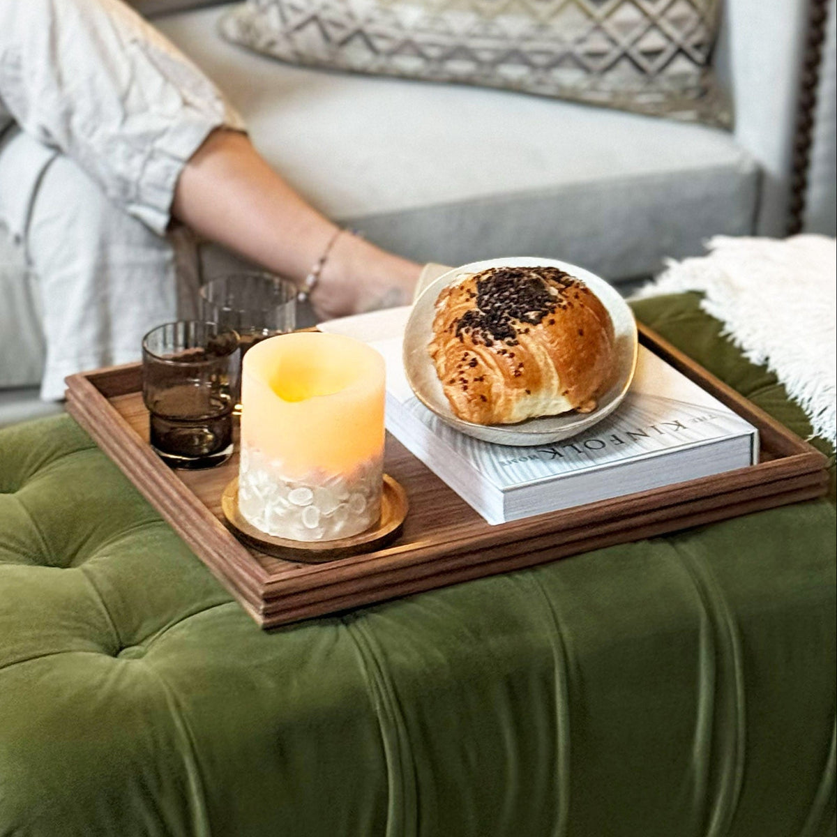 An olive green tufted velvet ottoman in a cosy living room setting, styled with a walnut tray holding a candle, a glass, and a croissant on top.