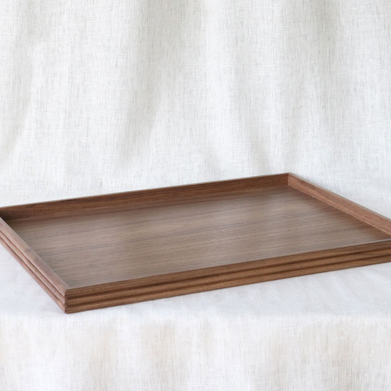 Angled view of an empty rectangular walnut wooden tray, showcasing its smooth surface and reeded edge detail, placed on a soft white linen background.