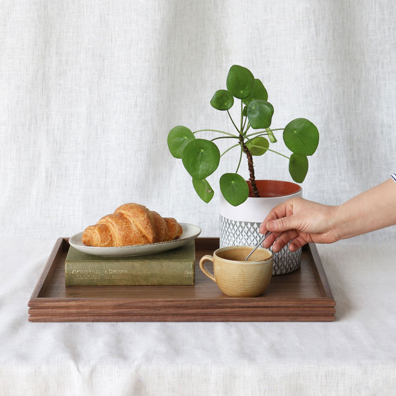 The tray arranged with a croissant on a book, a mug, and a potted plant, with a hand reaching in to stir the drink, showcasing its practical use.