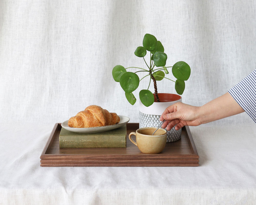The tray arranged with a croissant on a book, a mug, and a potted plant, with a hand reaching in to stir the drink, showcasing its practical use.