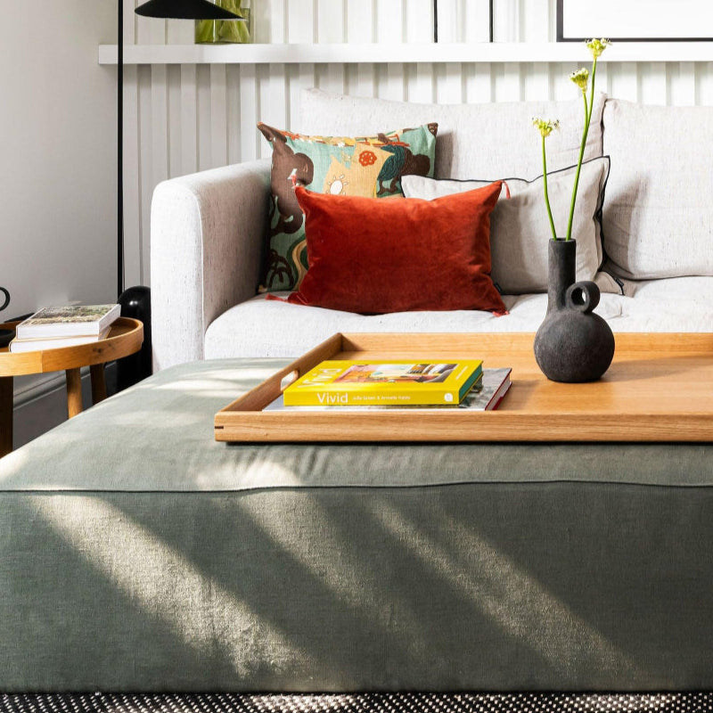 Large wooden ottoman tray placed on a green footstool in a cozy living room, featuring a yellow book and a sculptural black vase.