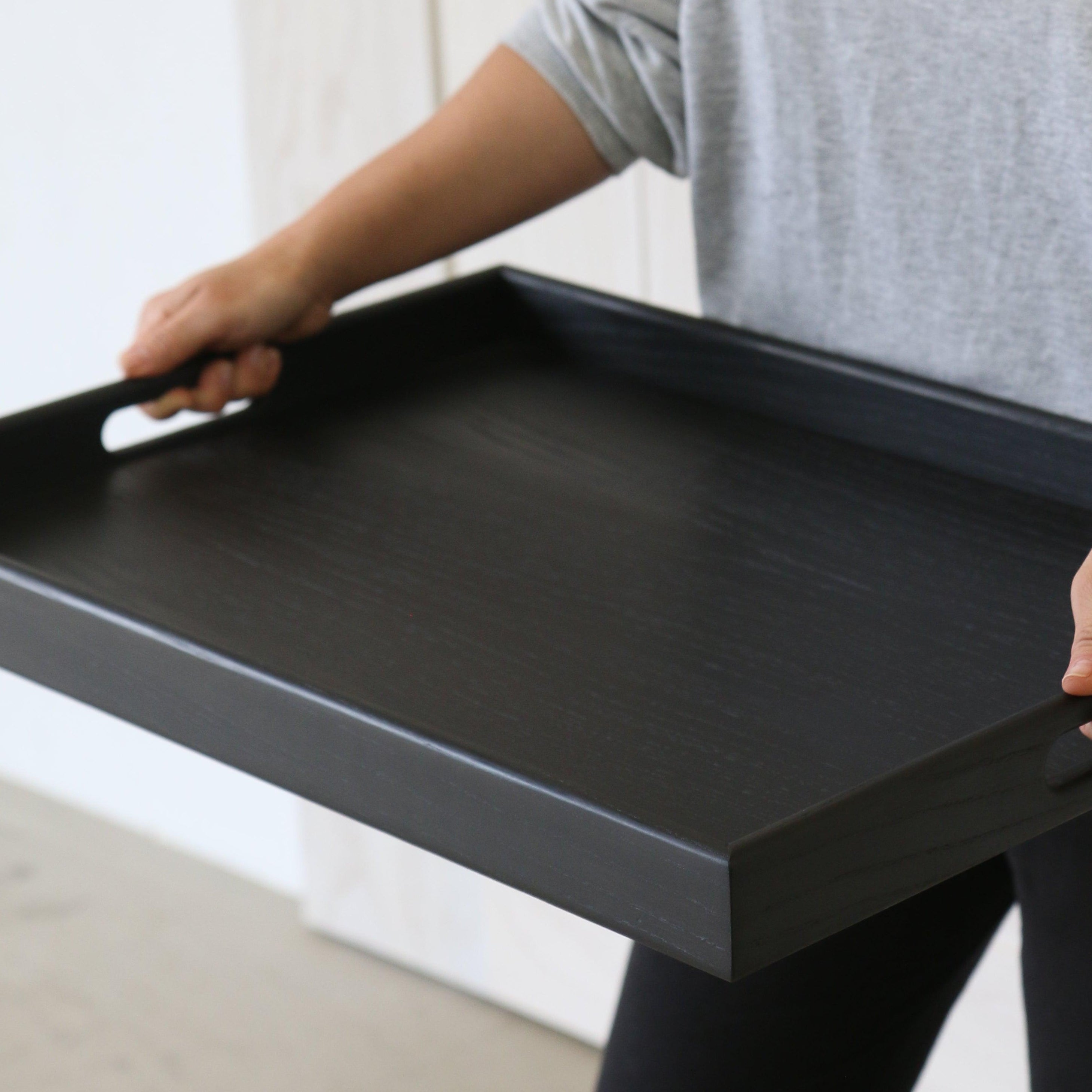 A wooden tray for footstool is carried by a person, showing the sleek black wood finish and integrated handle design.