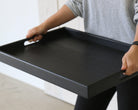 A wooden tray for footstool is carried by a person, showing the sleek black wood finish and integrated handle design.