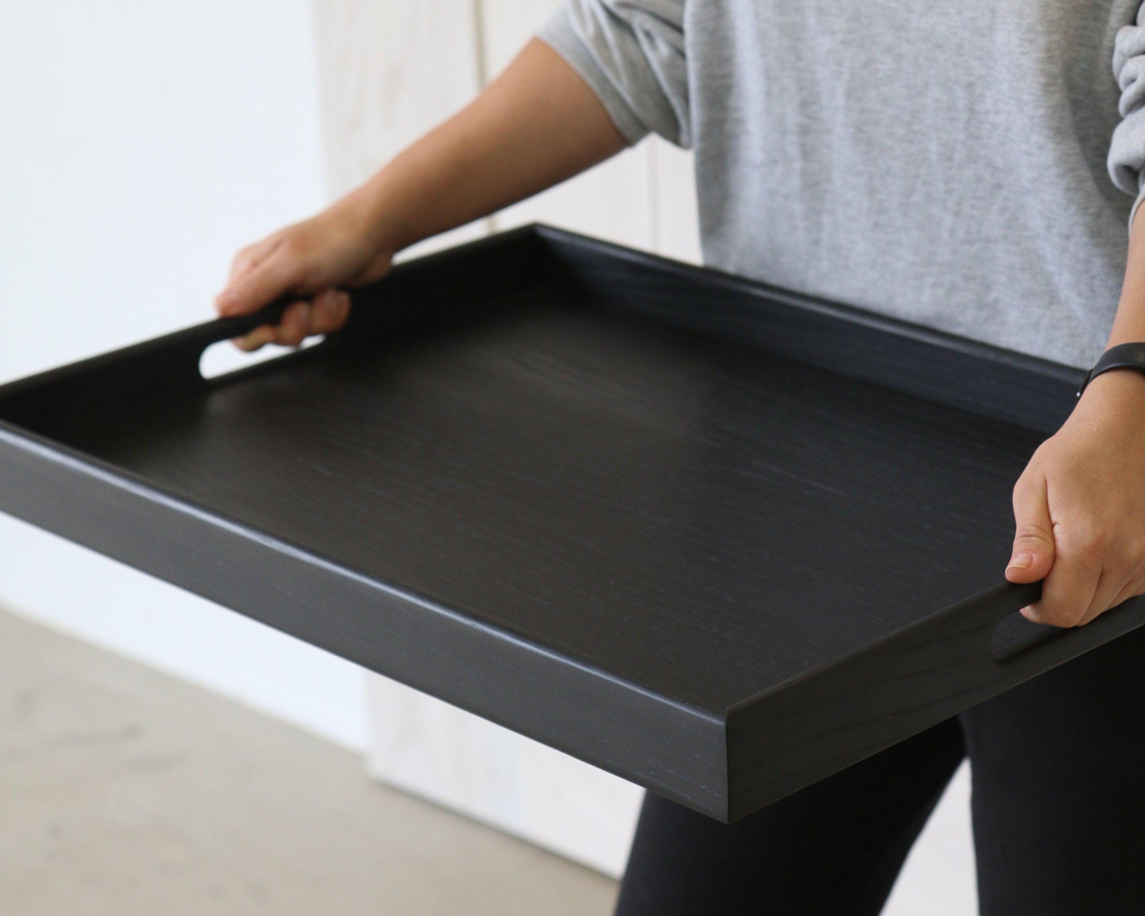 A wooden tray for footstool is carried by a person, showing the sleek black wood finish and integrated handle design.