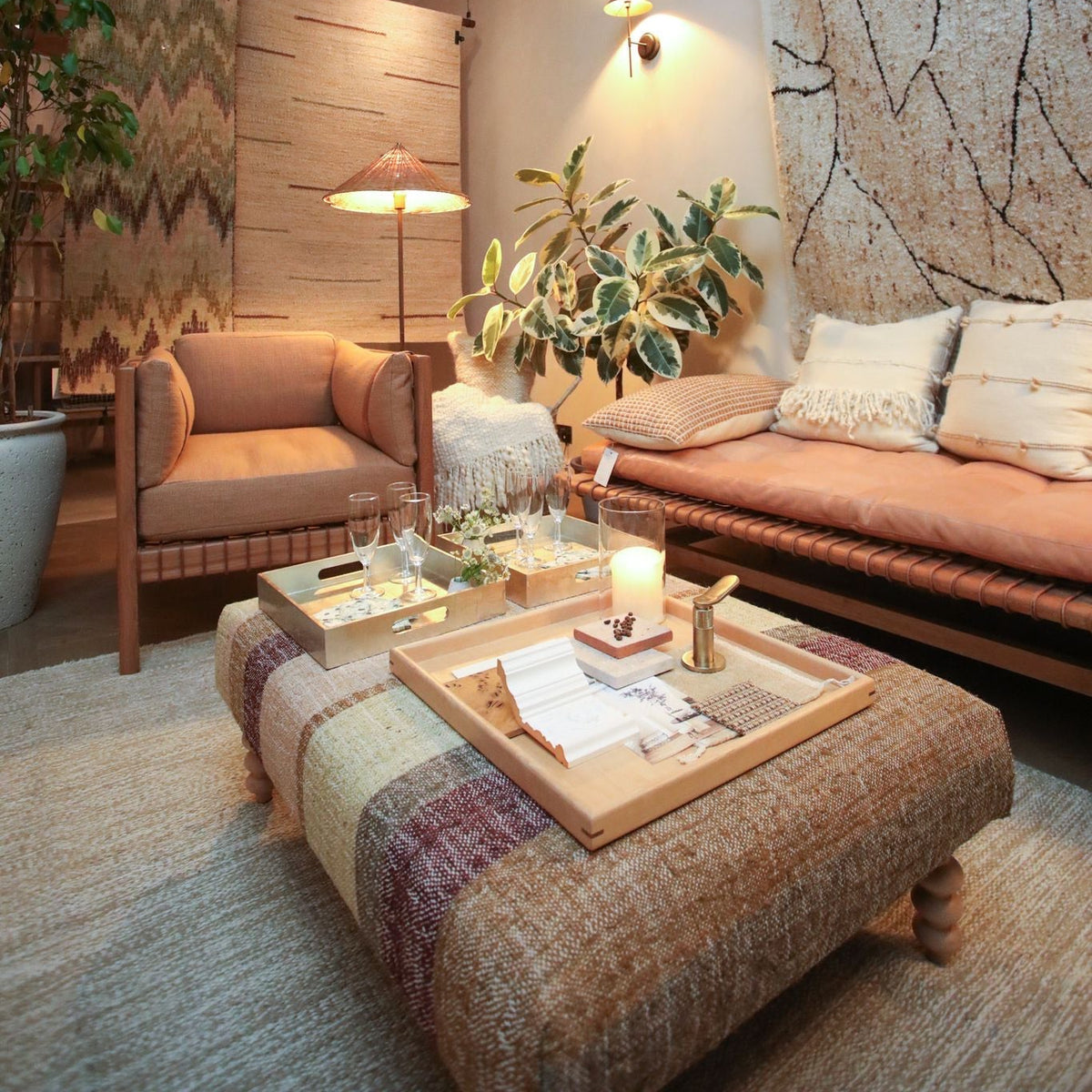 A cozy, rustic-style living room featuring a large woven ottoman in the foreground, a cushioned daybed with peach bedding and patterned pillows, a matching armchair, and a large abstract wall hanging under a slatted wood ceiling.