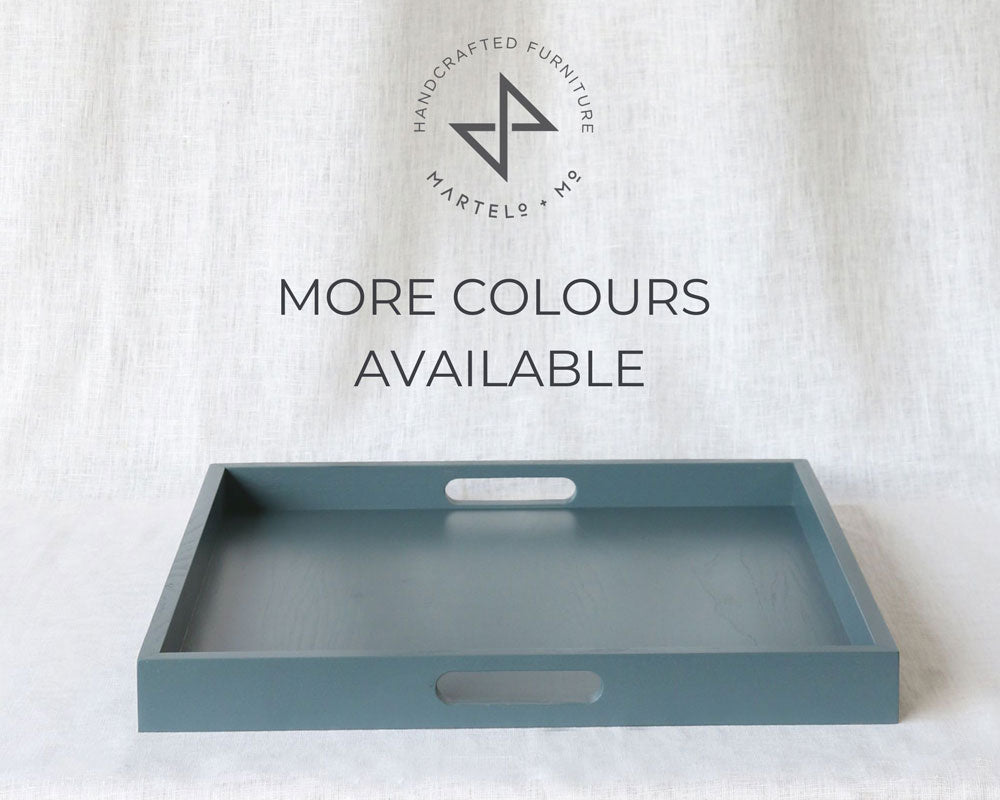 Blue-grey ottoman tray with cut-out handles and Martelo & Mo branding above, set against a light fabric backdrop with the text “More Colours Available.”