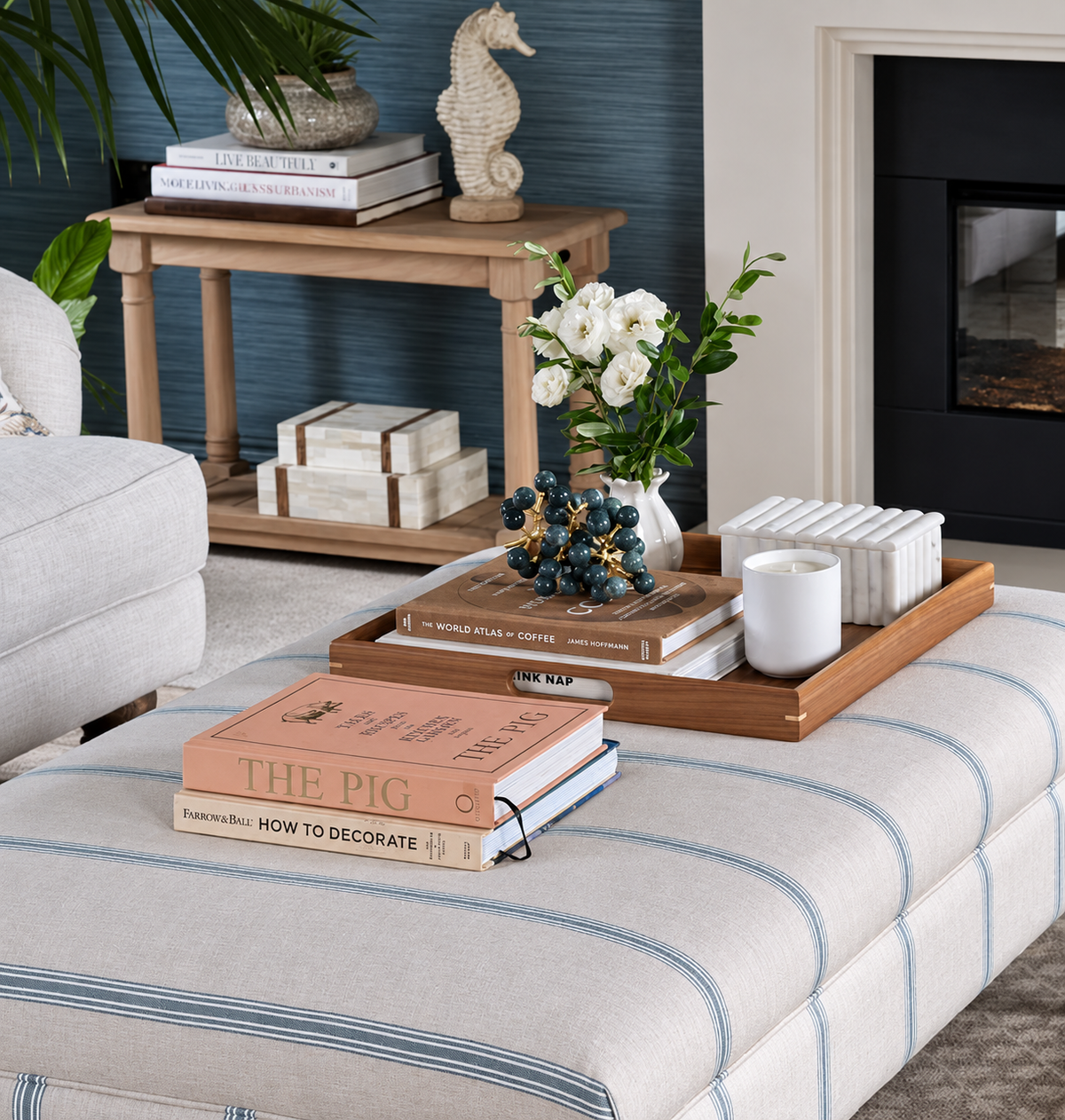 A styled living room scene featuring a large upholstered ottoman with blue pinstripes, topped with a walnut tray holding books, a candle, and a vase of white flowers. A matching wooden side table stands in the background against a dark blue panelled wall.