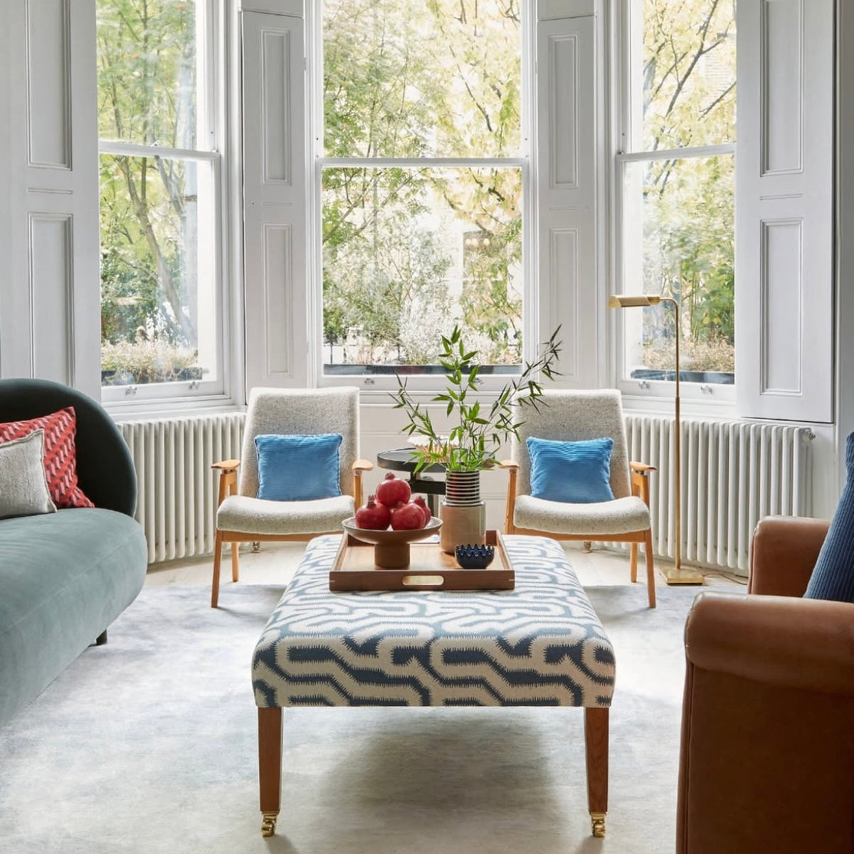 A bright, elegant living room with a large bay window looking out onto trees. The room is styled with a patterned blue and white coffee table, two cream armchairs with blue pillows, a green sofa, and a gold floor lamp.