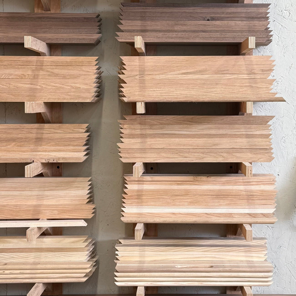 Stacks of natural wood planks displayed on wooden racks against a textured off-white wall, with a white electrical outlet visible on the right side.