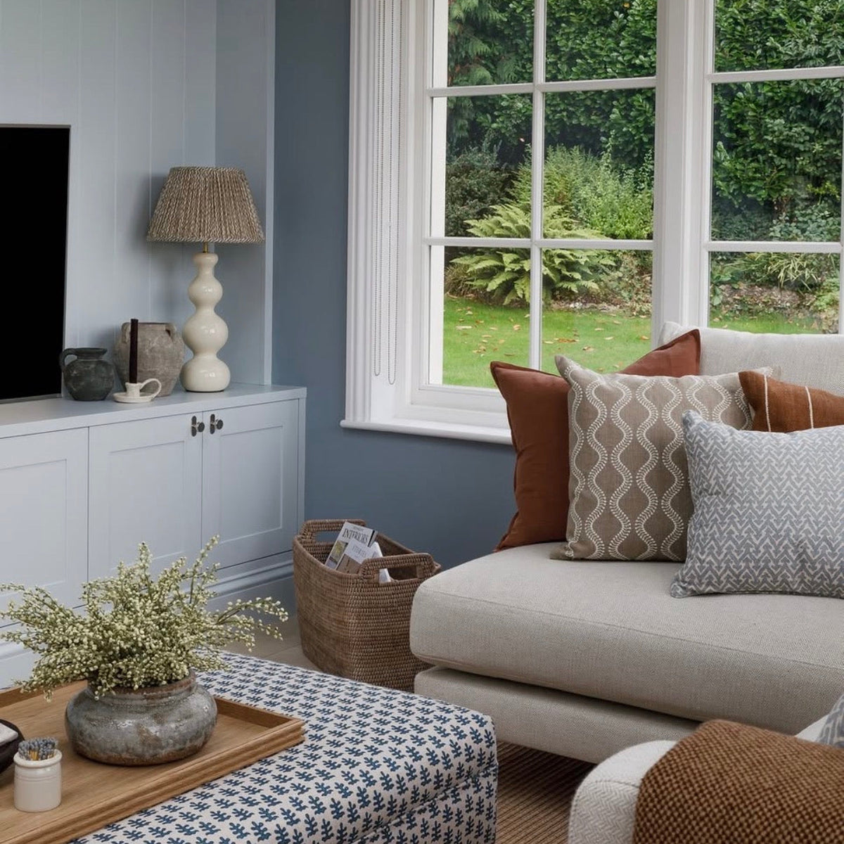 A styled living room corner featuring a light beige sofa with patterned and rust-colored throw pillows, a blue-and-white patterned coffee table, a white cabinet with a lamp, and a large multi-pane window looking out to a green garden.