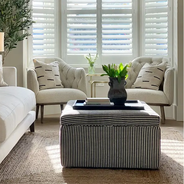 A bright, modern living room featuring white plantation shutters on a bay window, two white tufted armchairs with patterned pillows, a striped ottoman coffee table, an extra-large black ottoman tray, and a large potted green plant.