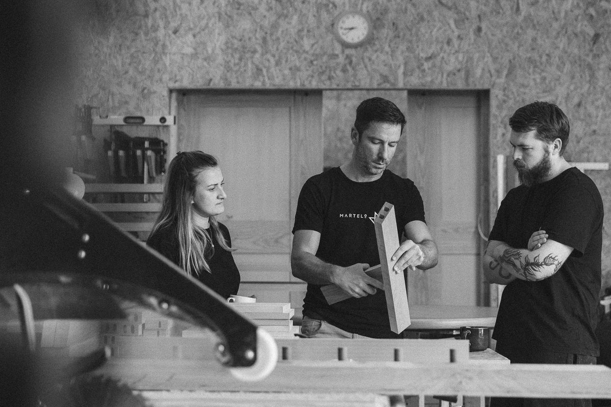 A black-and-white lifestyle photograph of three people in a workshop setting. The central figure, wearing a black t-shirt with the brand name 'MARTELO & MO' printed on the chest, holds a wooden board.