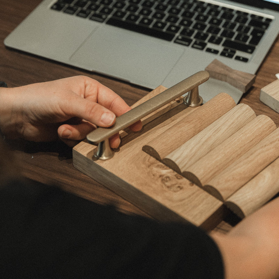 A person at a wooden desk holds a metal cabinet handle over a set of wood samples, with a laptop in the background.