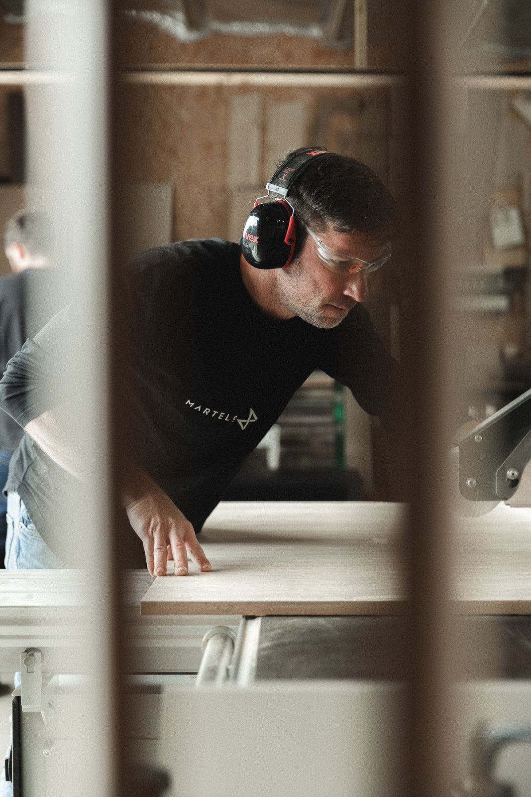 A man wearing a black long-sleeve shirt with a white 'MARTELO & MO' logo, safety glasses, and ear protection operates a table saw in a woodworking shop, cutting a light-colored wood board.