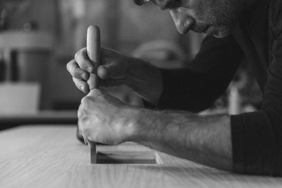 A black and white photograph of a craftsman's hands using a wooden chisel to carve or mark a piece of wood on a workbench.