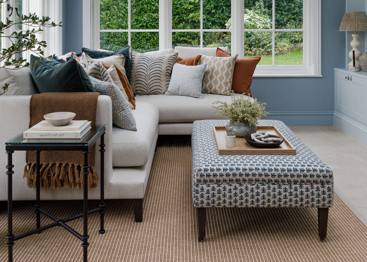 A bright, modern living room featuring a light grey L-shaped sectional sofa styled with a variety of patterned and solid throw pillows, a matching patterned ottoman coffee table and oak serving tray, and a black metal side table. The room has blue-gray painted walls, a beige woven rug, and large multi-pane windows looking out to green trees.
