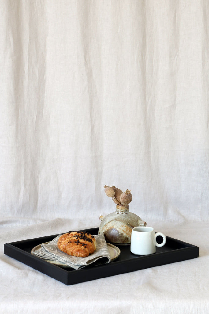 Medium-sized black wooden serving tray without handles with a chocolate croissant, espresso cup, and a vase.
