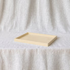 A minimalist rectangular tray in a light cream or pale yellow color, set against a draped, textured off-white fabric backdrop.