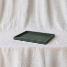 A dark green rectangular tray with a raised rim, displayed on a white textured fabric surface against a matching draped fabric backdrop.