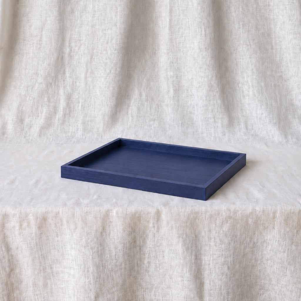 A rectangular indigo blue tray with raised edges, placed on a draped off-white fabric surface against a matching fabric backdrop.
