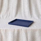 A rectangular indigo blue tray with raised edges, placed on a draped off-white fabric surface against a matching fabric backdrop.