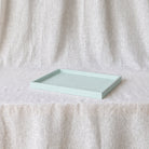A rectangular mint green tray with a shallow depth and raised edges, resting on a draped cream-colored fabric surface against a matching fabric backdrop.