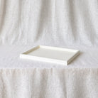 A minimalist rectangular tray with a raised rim, shown in a warm off-white or cream color, resting on a draped linen fabric backdrop.