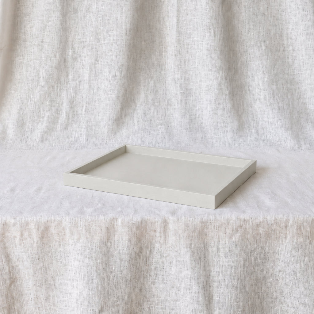 A minimalist rectangular tray with a raised rim, finished in a light warm off-white tone, displayed on a draped linen fabric backdrop.
