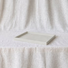 A minimalist rectangular tray with a raised rim, finished in a light warm off-white tone, displayed on a draped linen fabric backdrop.