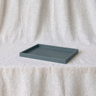 A rectangular matte smoke-blue tray with raised edges, placed on a draped light beige fabric surface against a matching fabric backdrop.