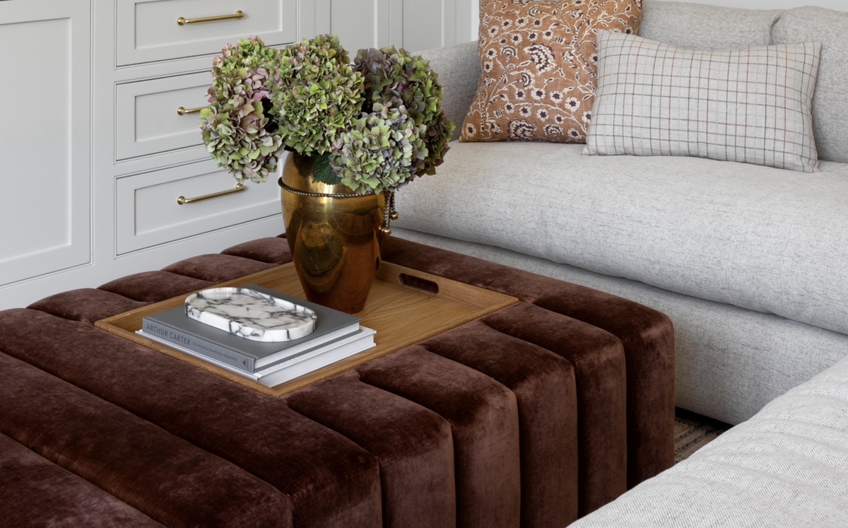 A brown velvet ottoman with a wooden tray top in a modern living room setting, styled with a bronze vase of green hydrangeas and books.